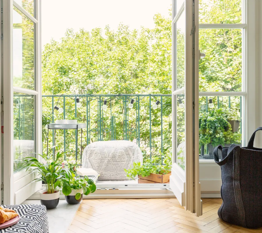 Öppen glasdörr från ett vardagsrum mot en balkong med gröna trädtoppar i bakgrunden.